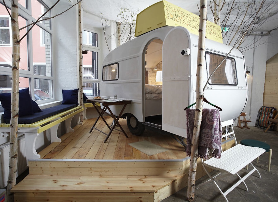 En liten vit husvagnstrailer installerad inomhus på en träplattform, designad som en inomhusglamping eller alternativt boende.